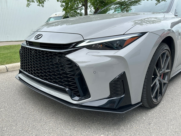 Close-up of the front end of a silver Lexus sedan parked outdoors on asphalt, showing the large black grille, headlights, and wheel.